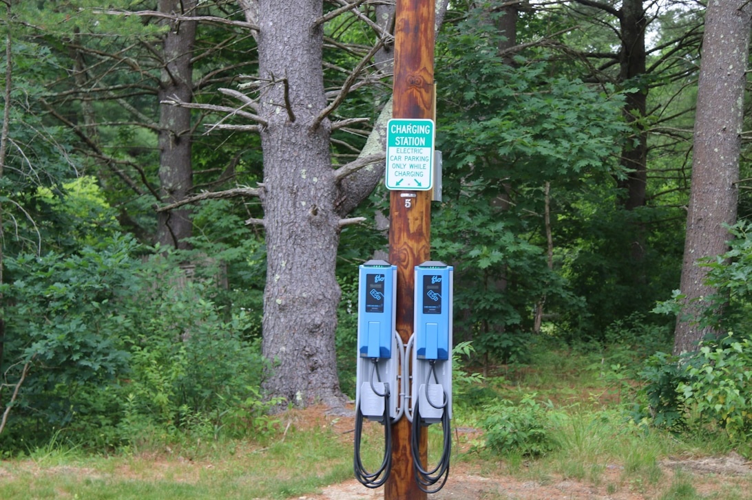 Två blå laddboxar monterade på en trästolpe i ett skogsområde, vilket visar en flexibel installationslösning utanför en traditionell husvägg.
